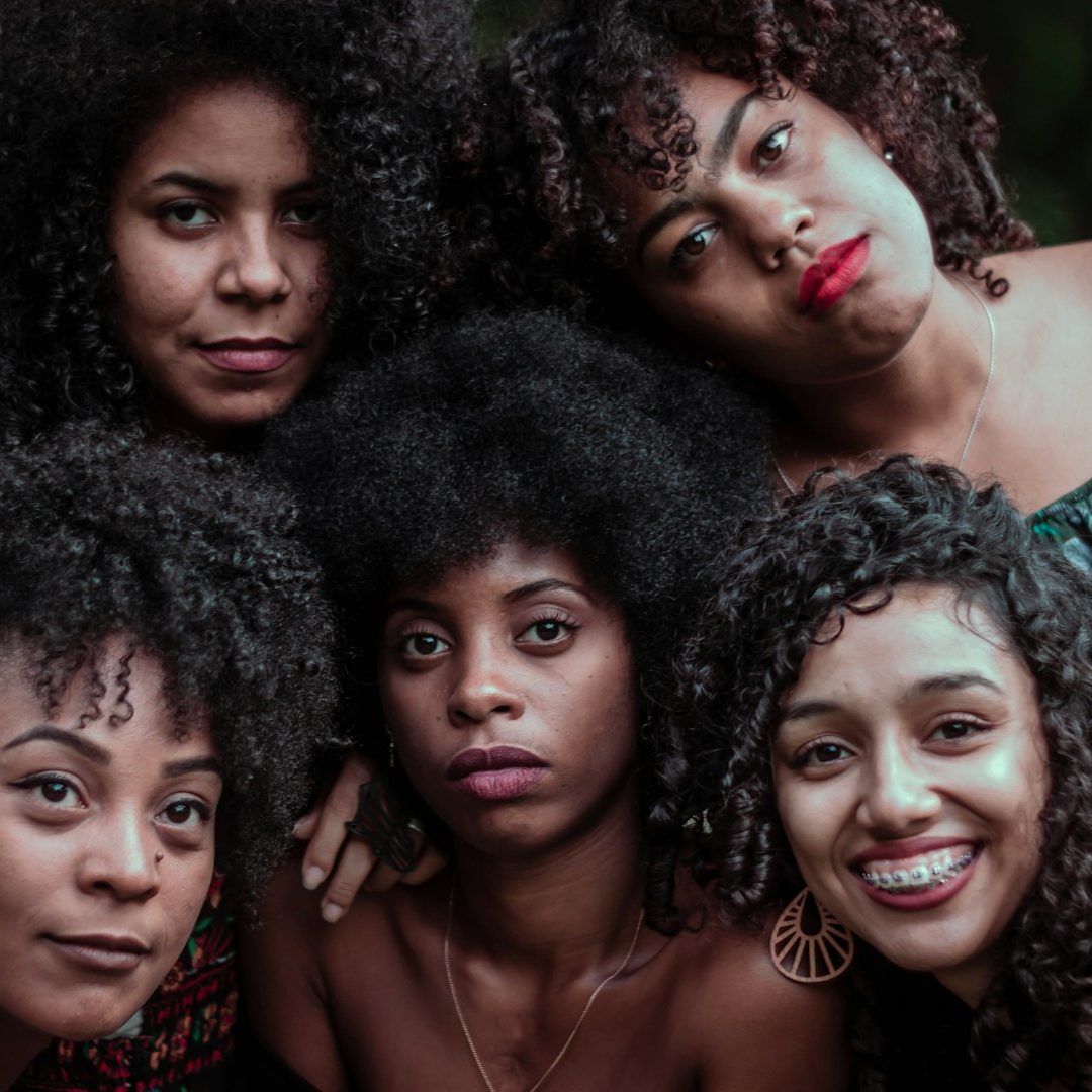 Five women of color with curly hair pose closely together, each displaying unique expressions.