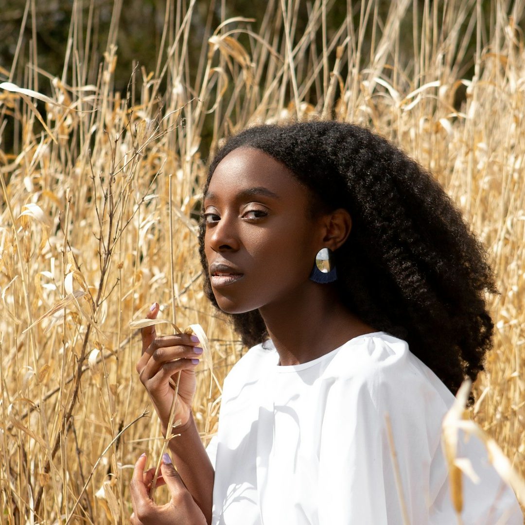A black woman with curly hair stands among tall golden grass, wearing a white blouse and earrings.